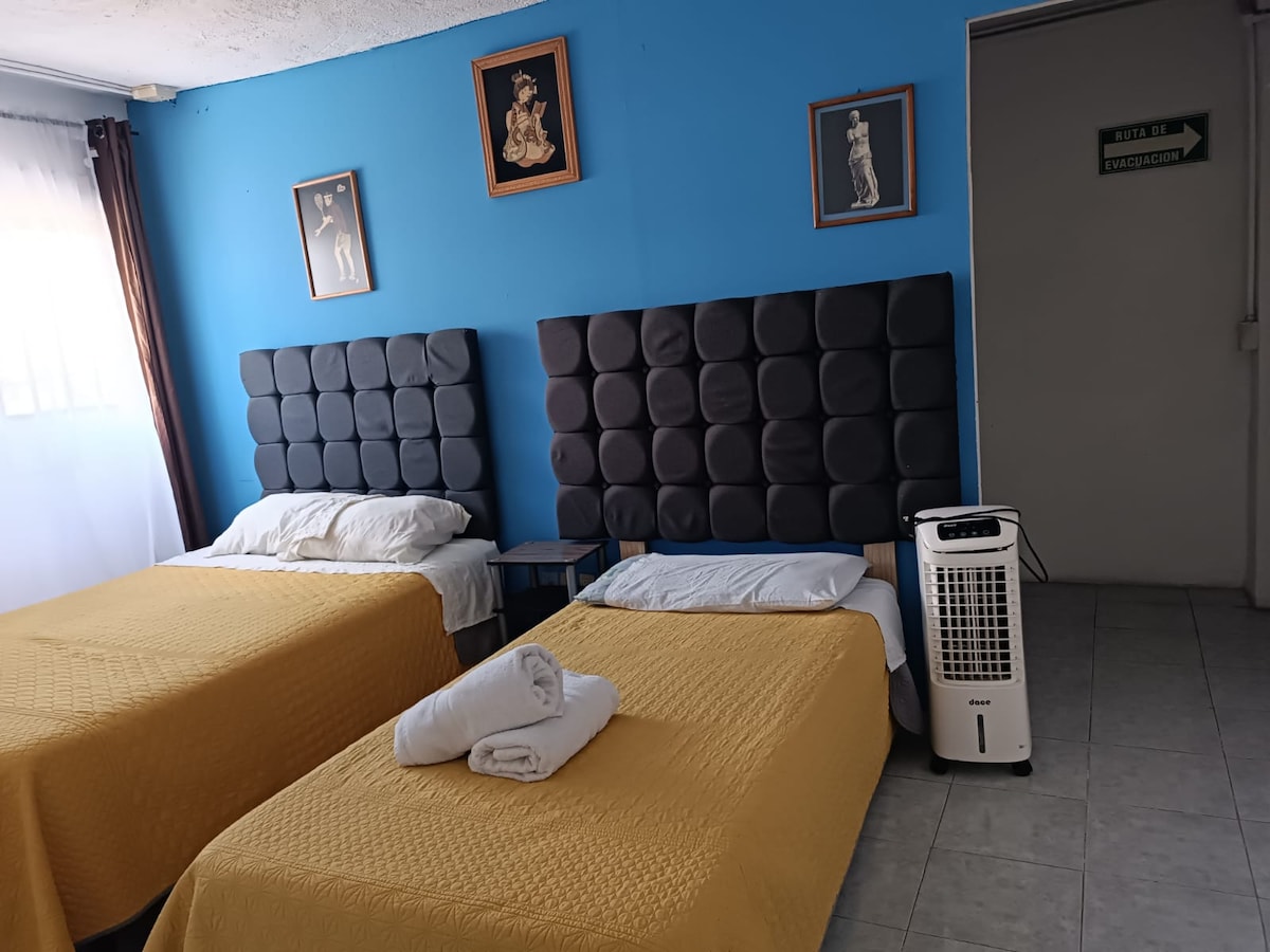 The room features two beds with dark upholstered headboards, each covered by a yellow bedspread. A white towel is neatly placed at the foot of one bed. Light enters through a window with sheer curtains, while decorative art pieces hang on the blue wall.