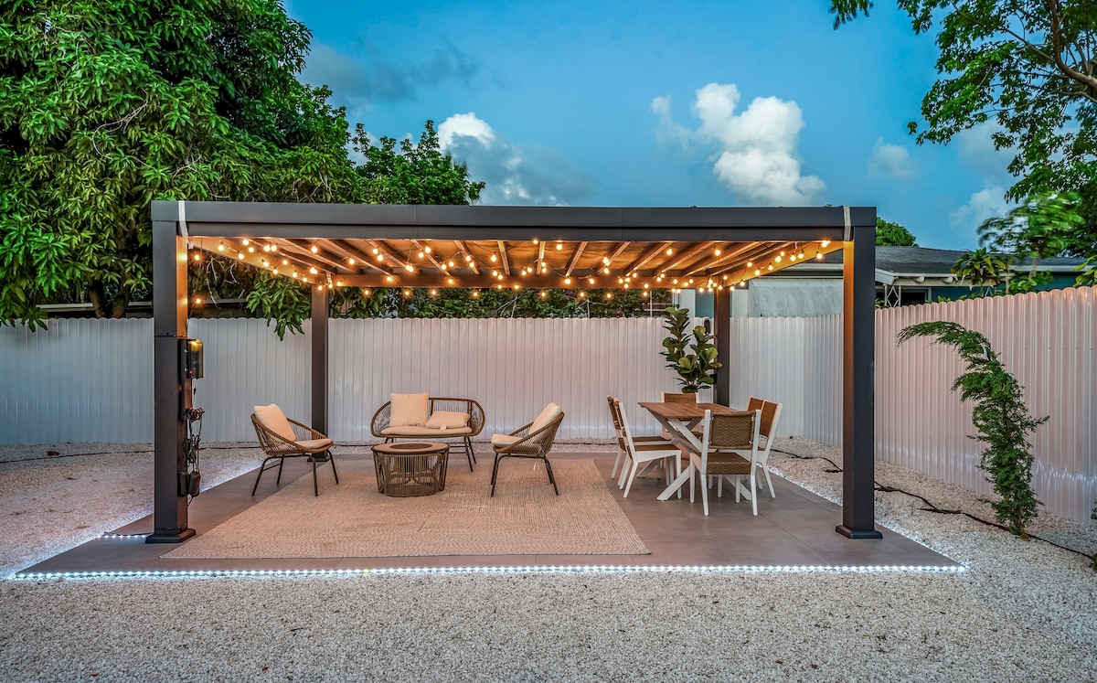 An outdoor pergola adorned with string lights provides an inviting space for relaxation and dining. Comfortable seating options include lounge chairs and a coffee table, complemented by a dining table surrounded by white chairs. The gravel floor adds texture to the serene environment.