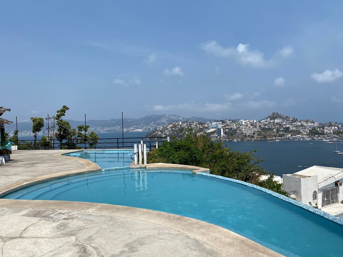 A serene swimming pool is seen overlooking the bay, with clear turquoise water reflecting the sky. Surrounding trees provide subtle greenery, and a scenic view of the coastal city unfolds in the background, showcasing the blend of nature and architecture.