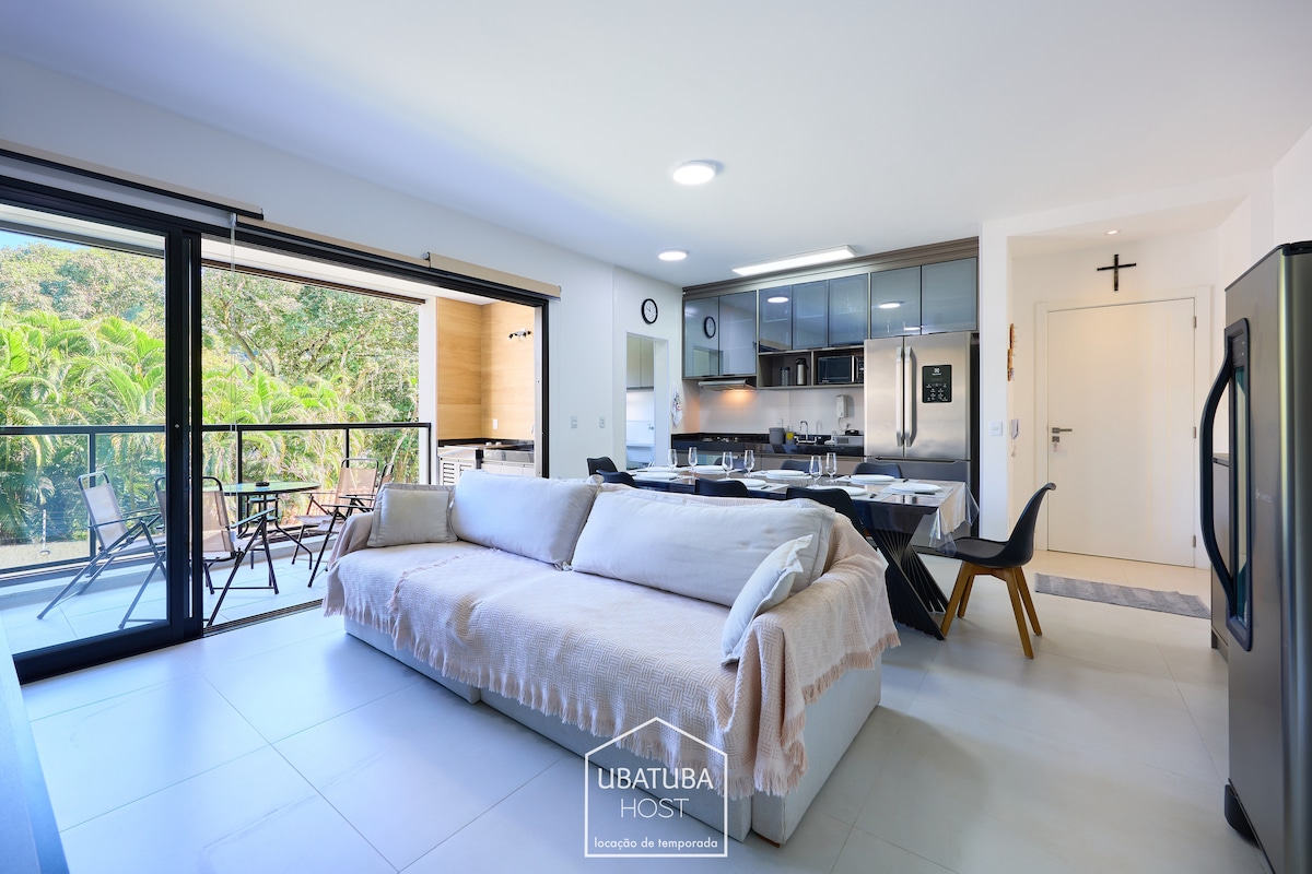 An open-concept living area features a modern sofa and a dining table set for six. Large sliding glass doors provide a view of the lush greenery outside, while a sleek kitchen is equipped with stainless steel appliances. Warm light illuminates the space.