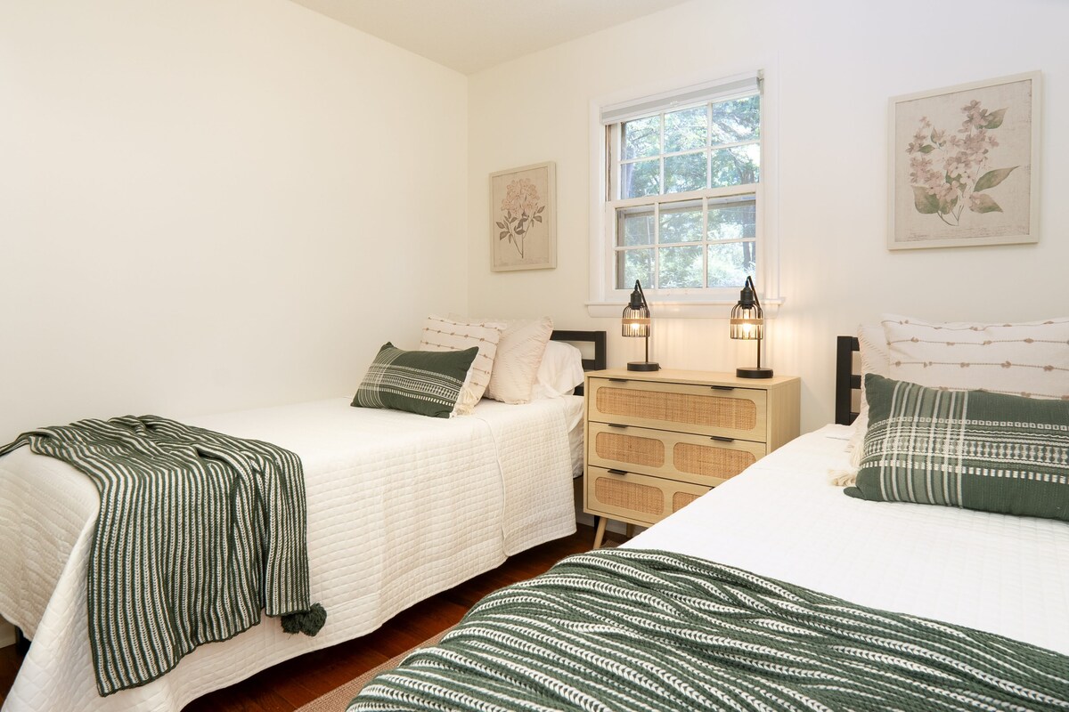 Šarmantna spavaća soba s bračnim krevetom i uređenjem inspiriranim prirodom. Bijeli kreveti s bočnim bočnim komodom od ratana i svjetiljkama za kavez. Zeleni naglasci i botanički otisci daju svježinu. S prozora se pruža pogled na vrt. Savršeno za dijeljenje ili dječju sobu u odmaralištu uz jezero.