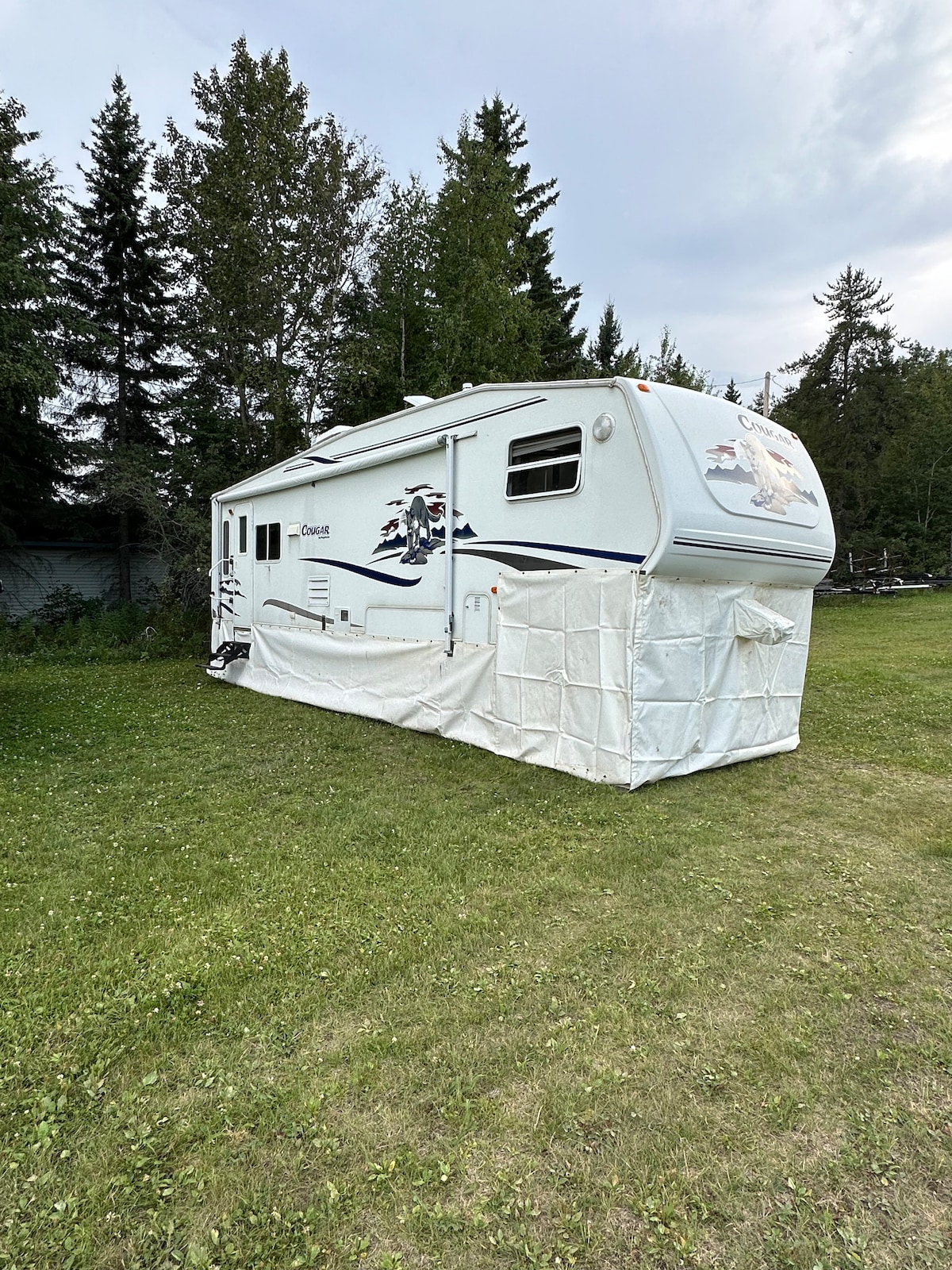 A white camper is positioned on a grassy area, surrounded by tall trees. The exterior features decorative elements, and a white protective cover is draped around the base, providing shelter. The setting offers a spacious outdoor space ideal for recreational activities.