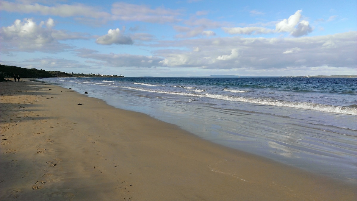 A wide shoreline extends along a tranquil beach, with gentle waves lapping at the sandy beach. The calm waters reflect a clear blue sky dotted with soft, white clouds, creating an inviting coastal scene. Lush greenery is visible in the background.