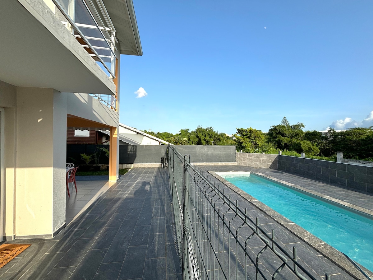 A private terrace is depicted, featuring a clear view of the pool. Sunlight reflects off the water, and a landscaped area surrounds the pool. The modern building's spacious exterior is visible, with a partial fence offering privacy and safety. The serene atmosphere is complemented by lush greenery in the background.