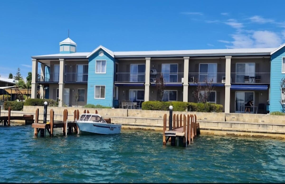 The building features a two-story design with blue exterior walls and a central turret. Balconies are present on the upper level, providing views of the water. A small boat is tied to a wooden jetty in the foreground, enhancing the waterfront ambiance.