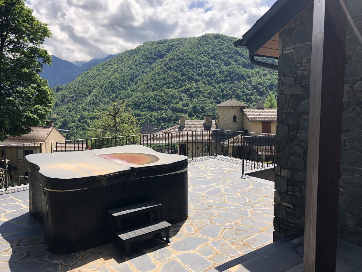 An outdoor area features a spa surrounded by a stone patio, with steps leading up to it. Lush green mountains rise in the background, and nearby buildings are visible, complemented by a partly cloudy sky.
