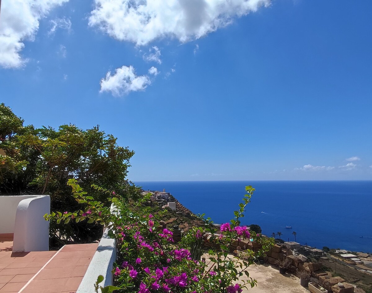 A view overlooking the sea showcases a bright blue sky scattered with clouds. Vibrant flowers add color to the foreground, framing the stunning ocean landscape. The scene reflects a tranquil coastal environment, inviting relaxation and admiration of the natural beauty.