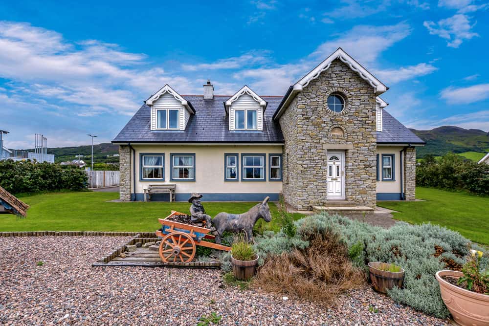 The exterior of a spacious house is presented, featuring a mix of stone and siding materials. A charming garden ornament depicting a person with a donkey is visible in the foreground, surrounded by greenery. Large windows reflect the bright sky above.