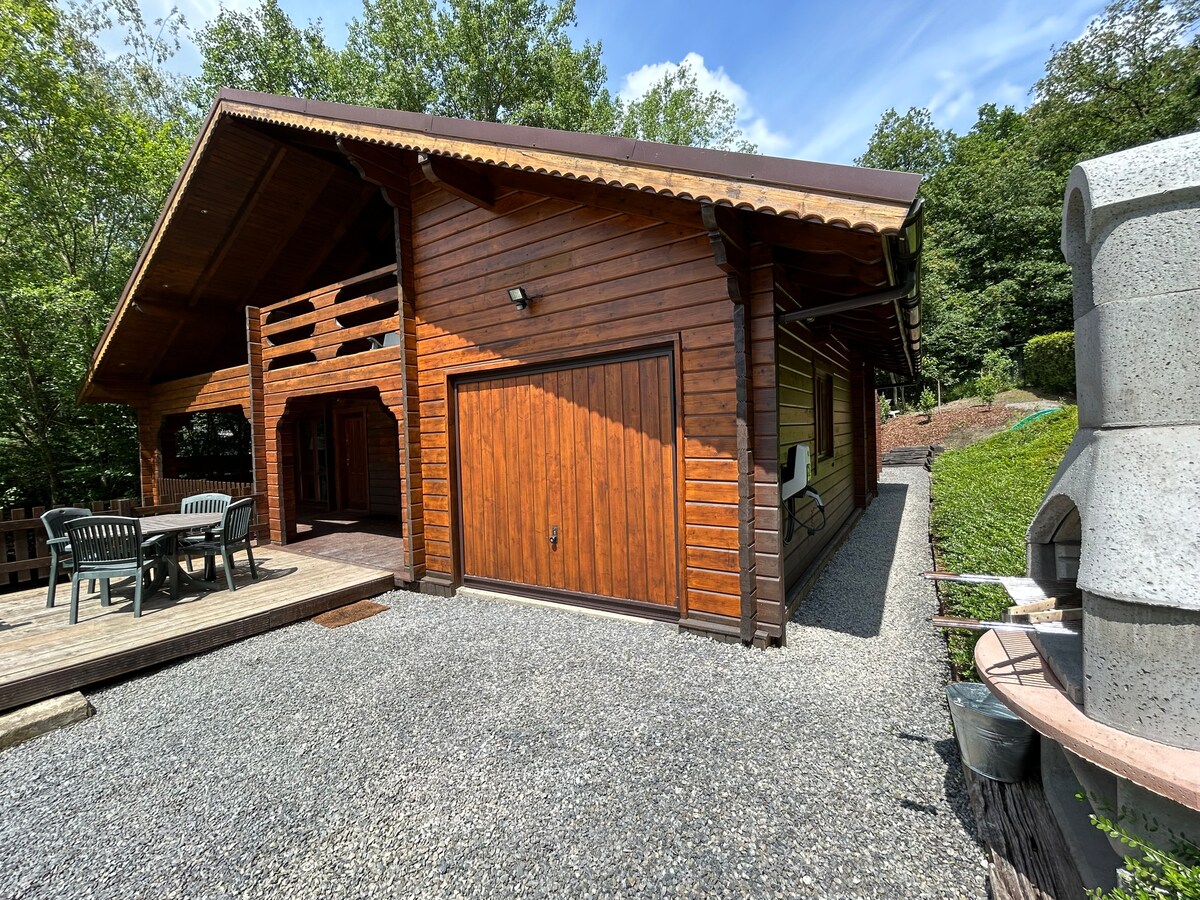 The exterior of a wooden chalet is captured, featuring a spacious deck with a dining set. A garage is seen on one side, while a wood-burning oven is located nearby. Lush greenery surrounds the property, providing a natural and serene setting.