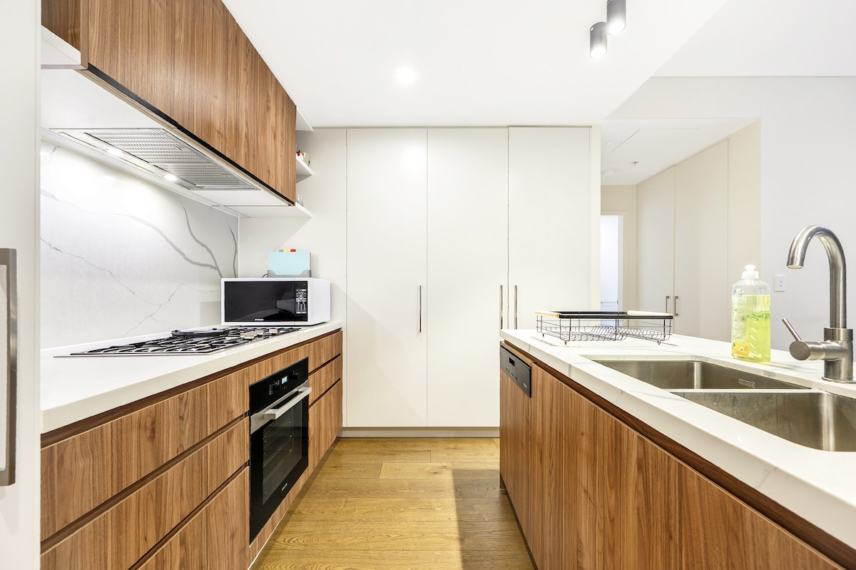 A modern kitchen is showcased, featuring sleek wooden cabinetry and white countertops. High-quality appliances, including a stove, oven, and microwave, are integrated into the design. Natural light illuminates the space, highlighting the spacious layout and contemporary finishes.