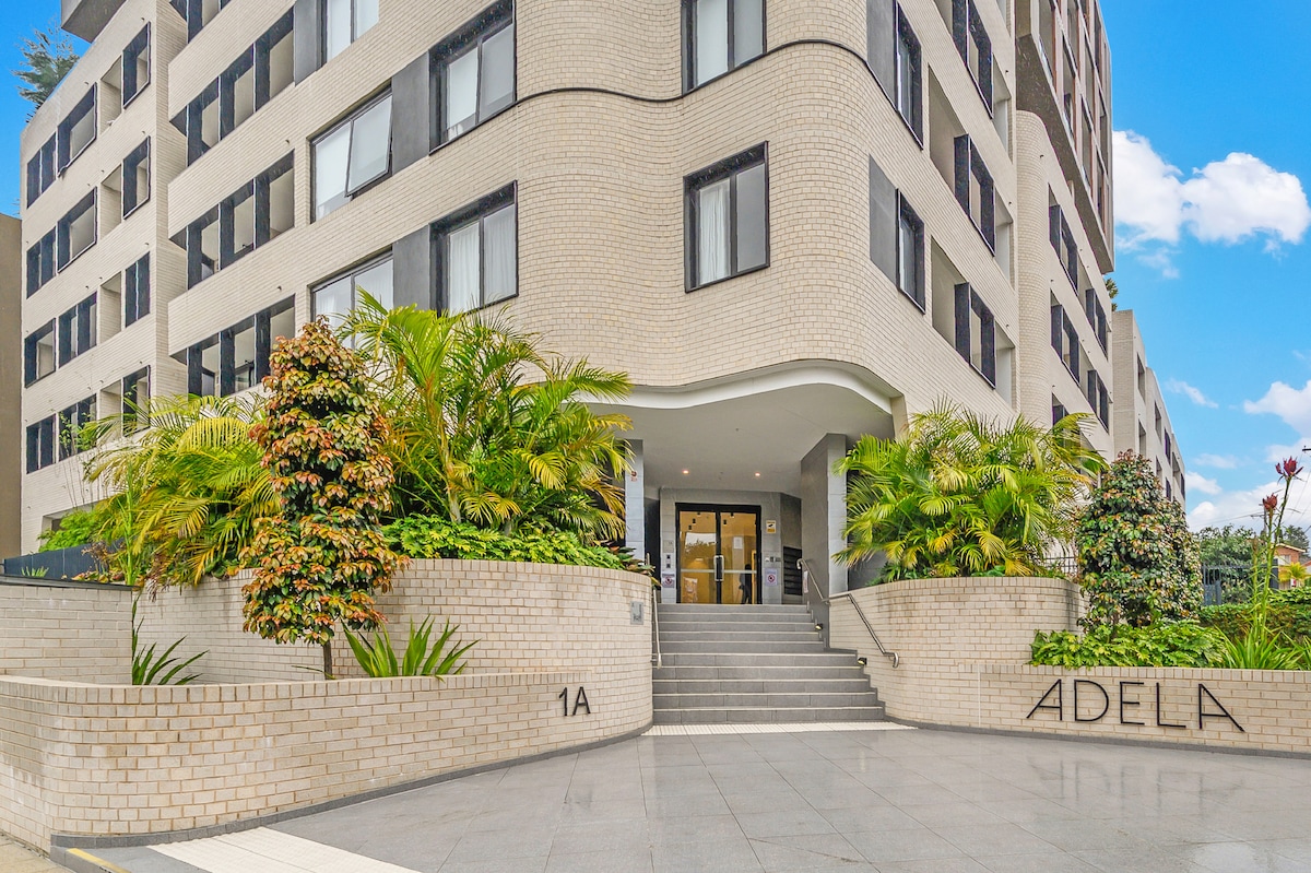 The entrance of the apartment building is framed by lush greenery and well-maintained landscaping. A set of stairs leads up to a welcoming lobby area. The building features modern architectural designs with large windows and a neutral brick facade.
