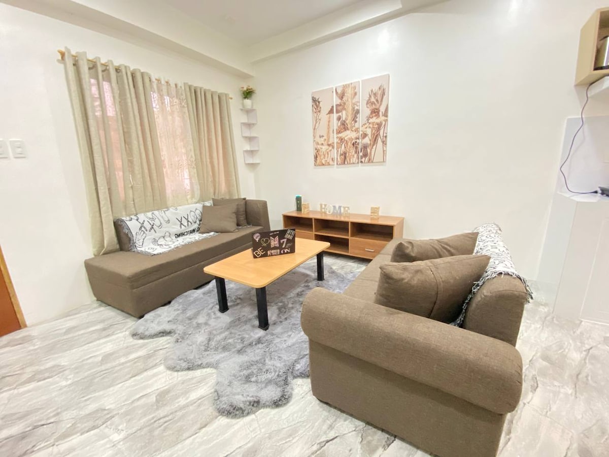 A spacious living room features a pair of cozy brown sectionals arranged around a wooden coffee table. Soft, textured rugs adorn the floor beneath, while natural light filters through light curtains. Simple wall art adds a touch of decor to the neutral-toned walls.