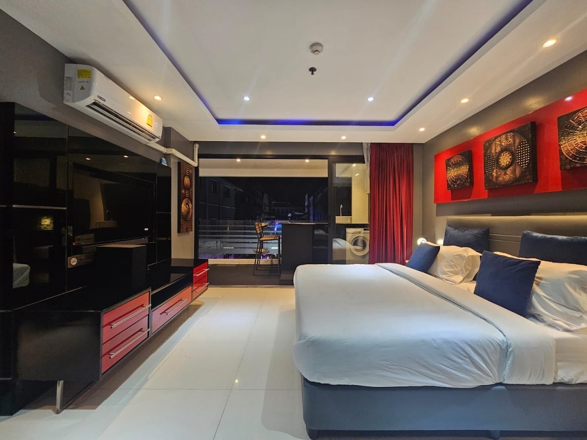 A modern guest room features a spacious bed with blue accent pillows. A sleek entertainment unit and bright red wall art enhance the decor. Natural light floods in through large windows, while a cozy seating area and kitchenette are visible in the background.