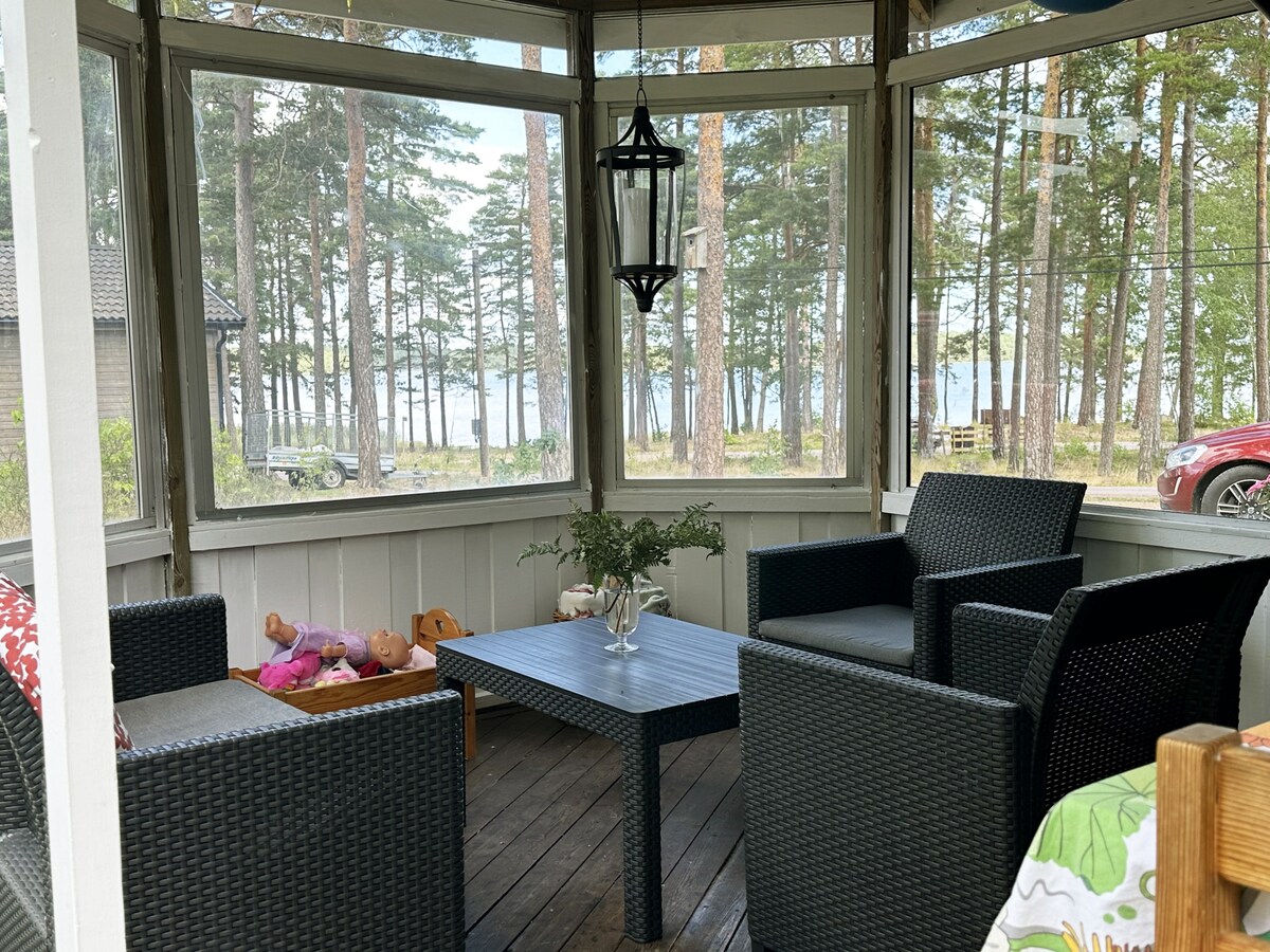 A screened-in porch features a wooden floor and a glass roof, allowing natural light to fill the space. Four dark wicker chairs surround a square coffee table, providing a cozy seating area. Trees and the water are visible in the background, enhancing the serene environment.