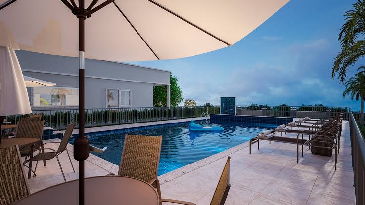 A spacious outdoor area features a large swimming pool surrounded by sun loungers and patio tables. Umbrellas provide shade for guests, and greenery frames the pool within a serene setting. The sky is filled with soft clouds, and the scene is illuminated by evening light.