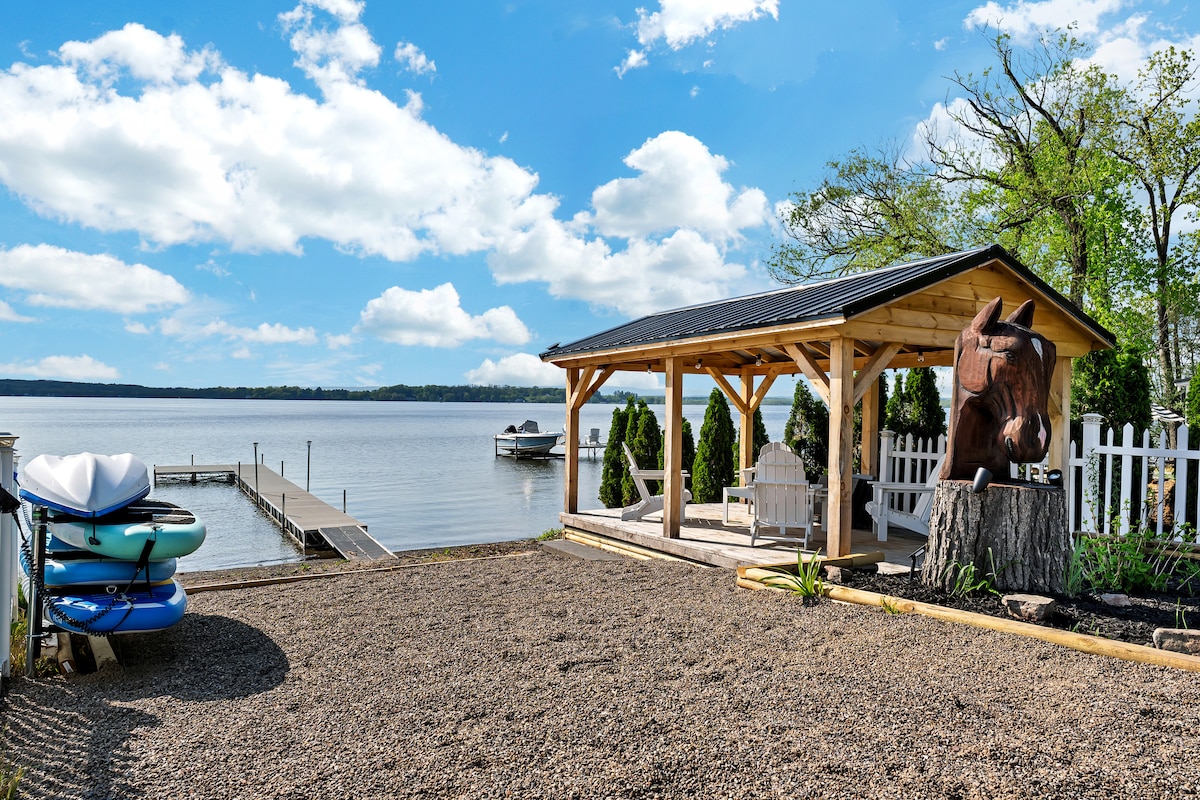 A serene lakeside view showcases a spacious wooden pavilion with a sculpture of a horse's head. Paddleboards are stacked nearby, alongside a boat dock extending into the calm water. Surrounding greenery and a white fence enhance the tranquil atmosphere of the outdoor space.