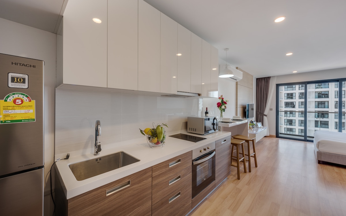 A modern kitchen is presented with sleek cabinetry and integrated appliances. A spacious countertop features a sink and stove, complemented by a small breakfast bar with seating. Natural light filters in from large windows, offering a view of the city outside.