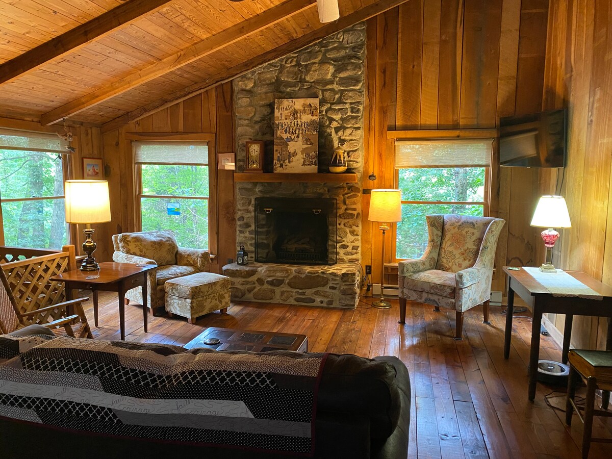 A warm living area is highlighted by a stone fireplace and wooden beams overhead. Two comfortable chairs are positioned near large windows, allowing natural light to illuminate the space. A cozy couch and side tables enhance the inviting atmosphere, complemented by lamps casting a soft glow.