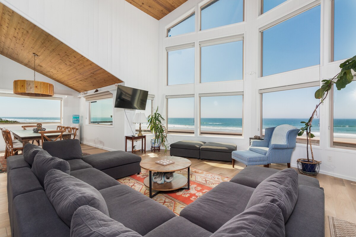 A spacious living room features large windows offering uninterrupted views of the Pacific Ocean. A comfortable sectional sofa, a coffee table, and a decorative rug are arranged around a central area. A cozy accent chair and a dining table can be seen in the background, along with a plant.