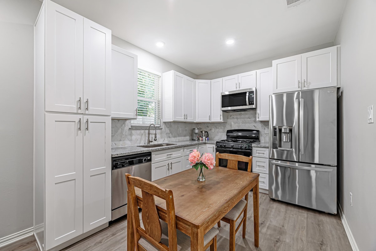 The kitchen features modern white cabinetry and stainless-steel appliances, including a refrigerator and gas stove. A wooden dining table is set for four, complemented by floral decor. Natural light enters through a nearby window, highlighting the sleek countertops and tile backsplash.