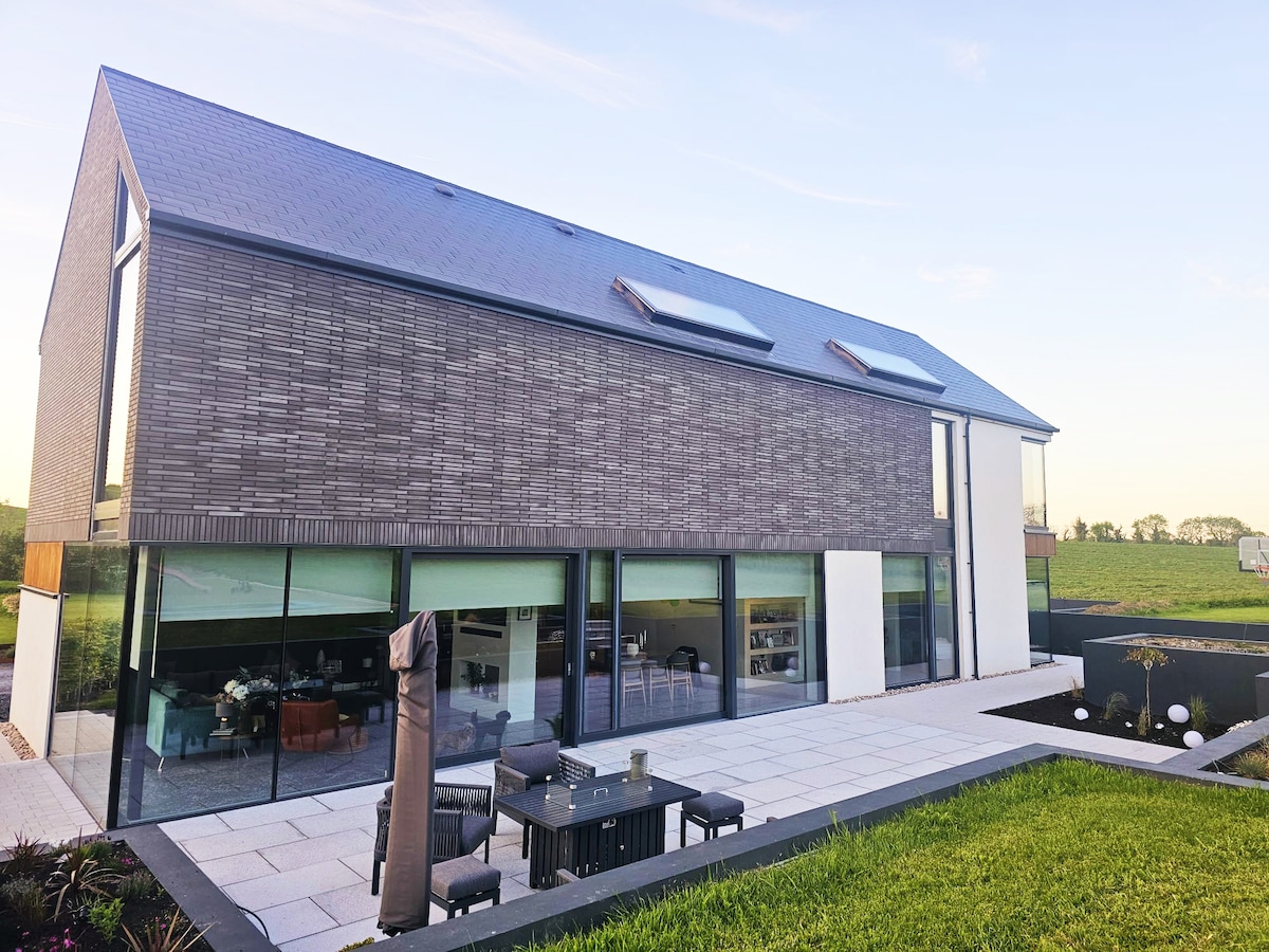 A modern two-story home is showcased, featuring a sleek facade of large windows and textured panels. The outdoor area includes a spacious patio with seating, set against a backdrop of landscaped greenery, creating a seamless connection with the surrounding countryside.