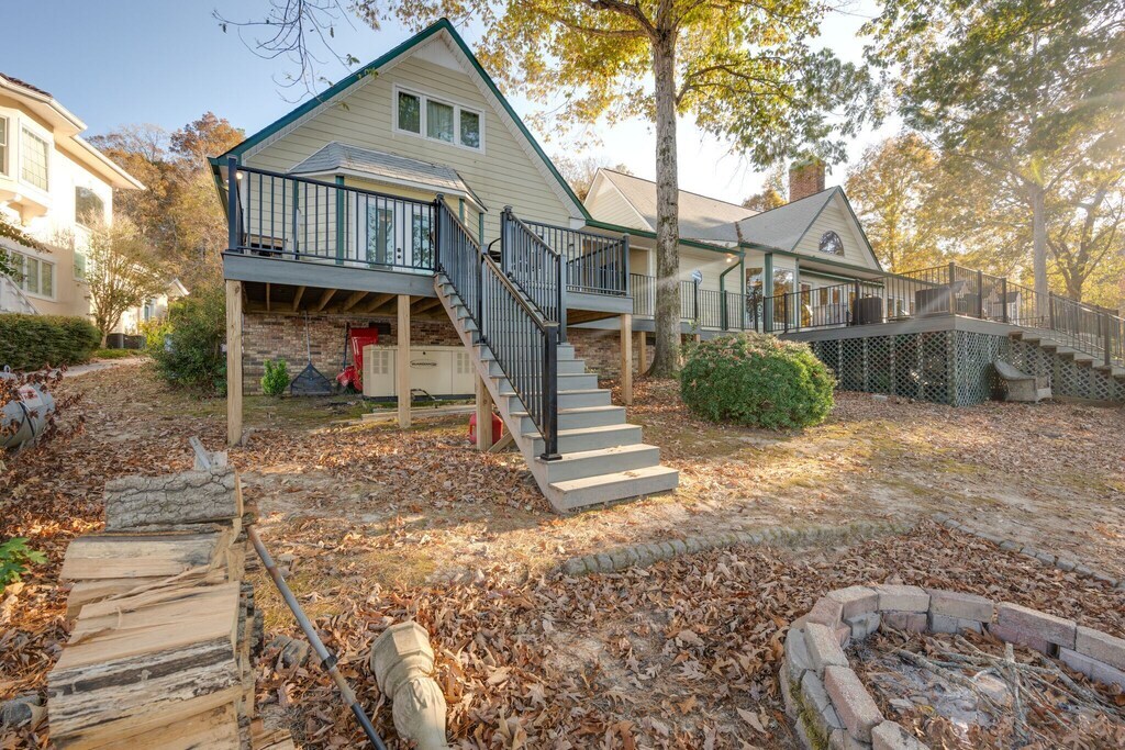 The exterior view showcases a two-story house with a spacious deck accessed by steps. The ground is covered in fallen leaves, with a fire pit made of stone visible in the foreground. Landscaping features shrubs around the base of the house.