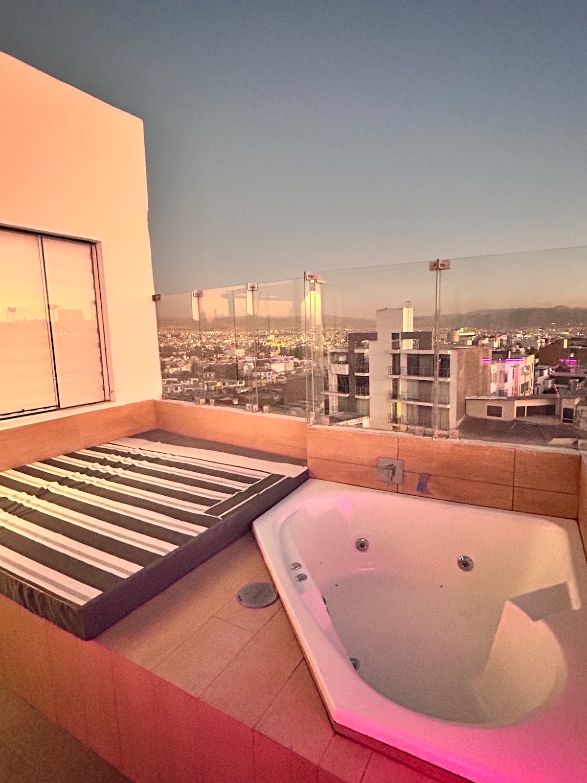 An outdoor terrace features a cozy seating area with a cushioned bench and a jacuzzi. The view overlooks the city, with lights visible in the distance as dusk settles. Surrounding glass railings provide a clear perspective of the vibrant atmosphere below.