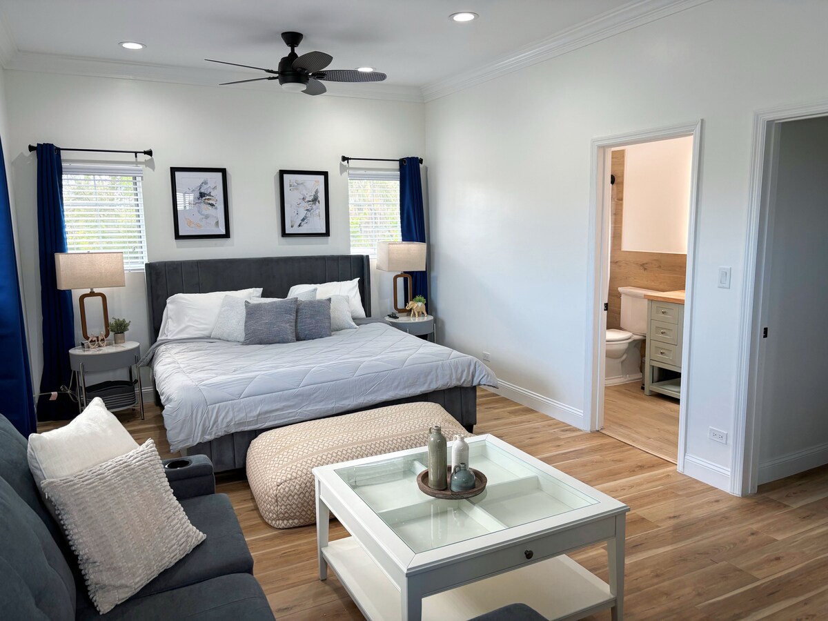 A spacious bedroom features a large bed with neutral bedding, complemented by decorative pillows. Bedside tables with lamps are positioned on either side. A comfortable seating area includes a sofa and an ottoman, while an ensuite bathroom is visible through an open door.