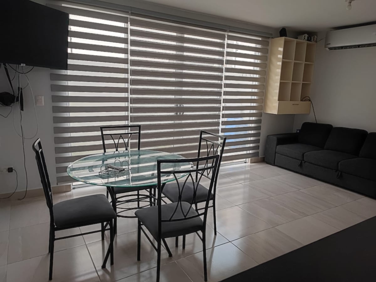 A dining area is seen with a circular glass table surrounded by four black chairs. A gray sofa is positioned against the wall, with shelves above. Light filters through horizontal blinds, creating a softly lit and open atmosphere.