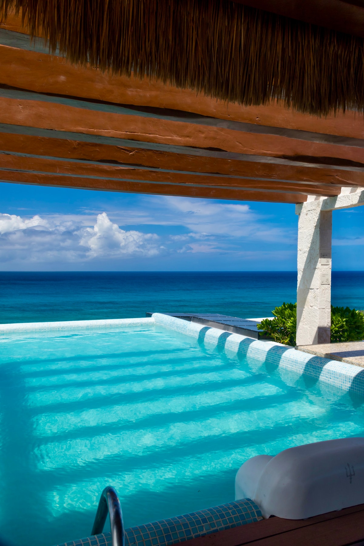 A refreshing swimming pool is framed by a thatched roof, with gentle waves visible in the background. The bright blue water mirrors the expansive Caribbean sea, while the clear sky is adorned with soft clouds, offering a tranquil setting.