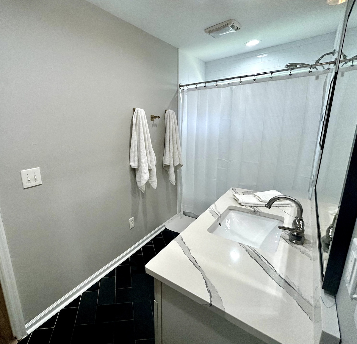 A clean bathroom features modern fixtures and a sleek countertop with a rectangular sink. Light-colored walls contrast with dark tiled floors. Two white bathrobes hang neatly on hooks. A shower is enclosed with a translucent curtain, providing privacy while maintaining an open feel.