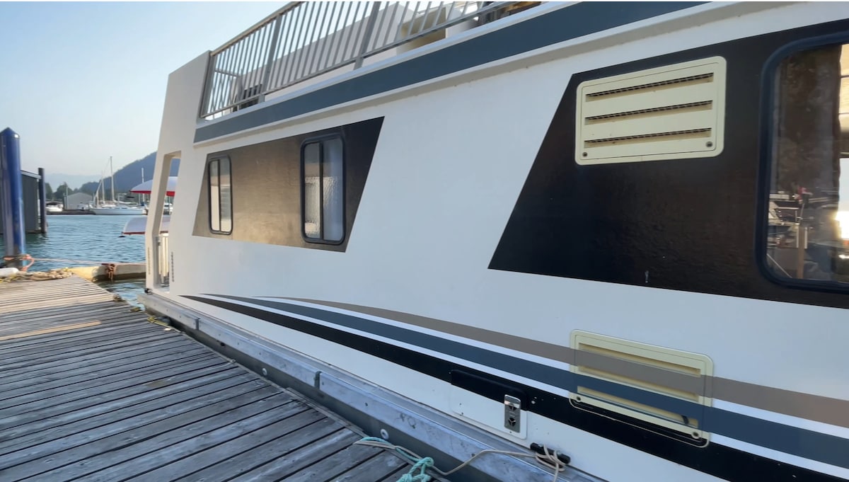 The exterior of the houseboat is visible, showcasing a sleek design with a combination of dark and light colors. The boat is docked, with wooden planks leading to the marina. The surrounding water reflects the serene environment, enhancing the connection to nature.