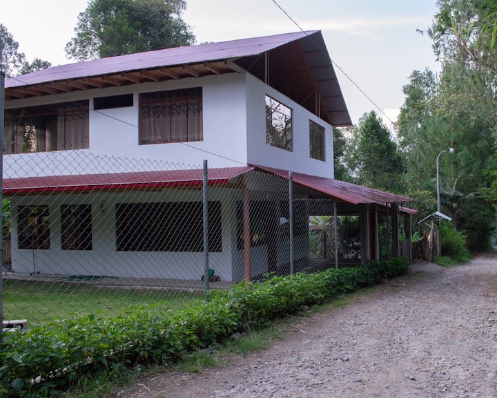 A two-story house is surrounded by greenery, featuring a red-roofed overhang and large windows. A gravel path leads to the entrance, and a fence is seen along the perimeter, offering a sense of privacy and seclusion.