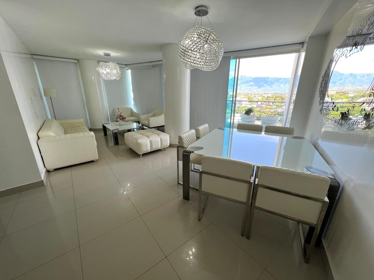 A bright living area features contemporary furniture, including a white sectional sofa and a glass dining table. Large windows provide a view of the surrounding landscape. Modern light fixtures hang from the ceiling, adding a touch of elegance to the space.