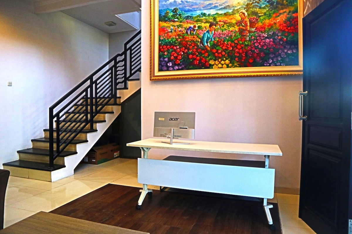 A workspace area features a sleek white desk equipped with a computer monitor. A vibrant floral painting adorns the wall above, while a staircase with a black railing leads to an upper level. Light reflects off the polished floor tiles.