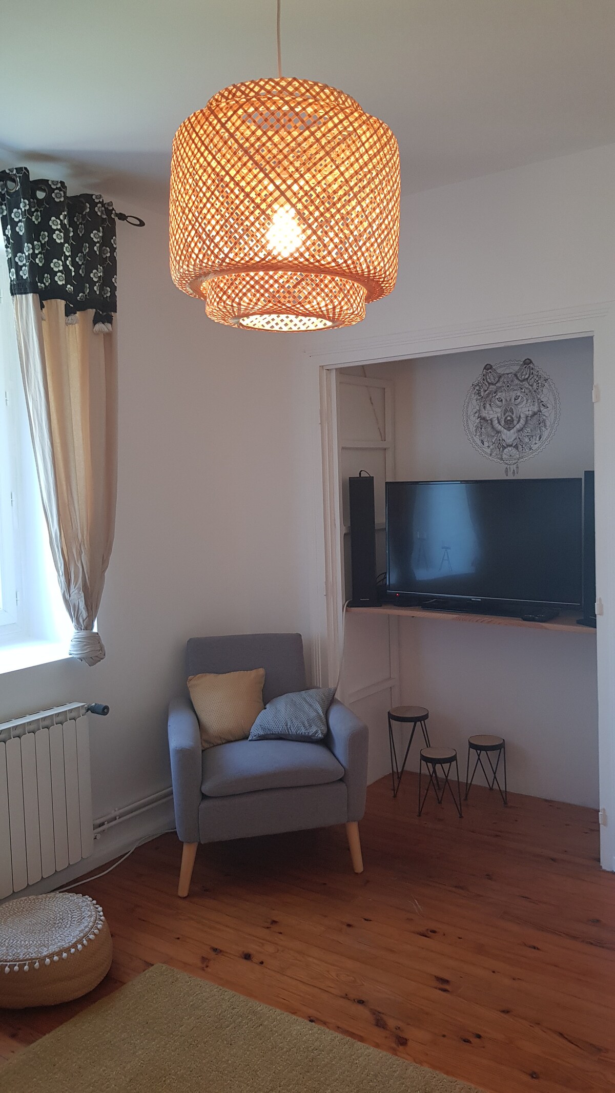 A cozy corner of the living area is highlighted by a woven pendant light above a comfortable armchair with a decorative pillow. A television is mounted on the wall, and two small side tables are positioned nearby. Natural light streams through the window with patterned curtains.