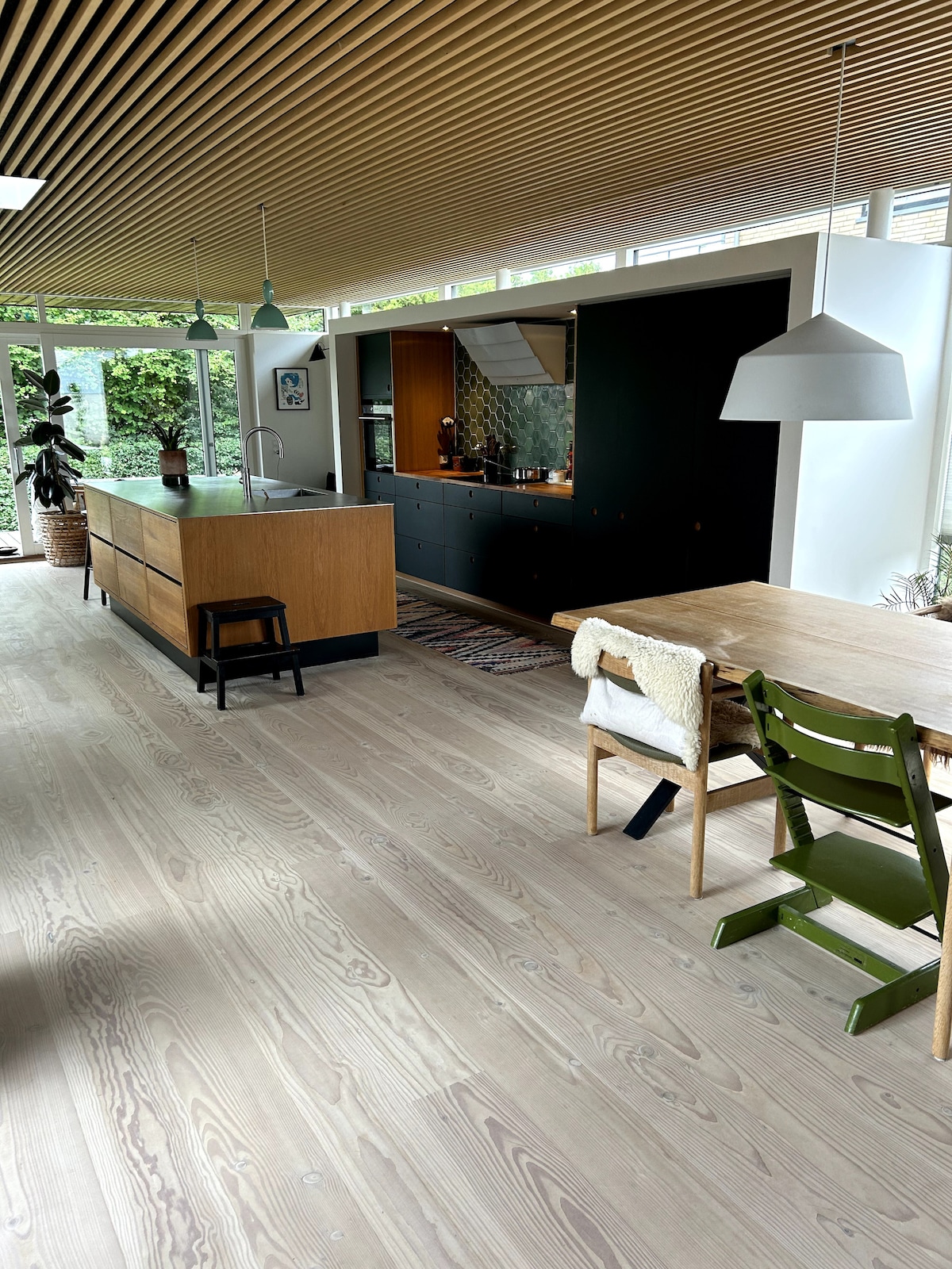 An open kitchen is highlighted by a center island with wooden cabinetry and green accents. A wooden dining table is set to one side, complemented by a green chair and a cozy throw. Abundant natural light fills the area, creating a welcoming space.
