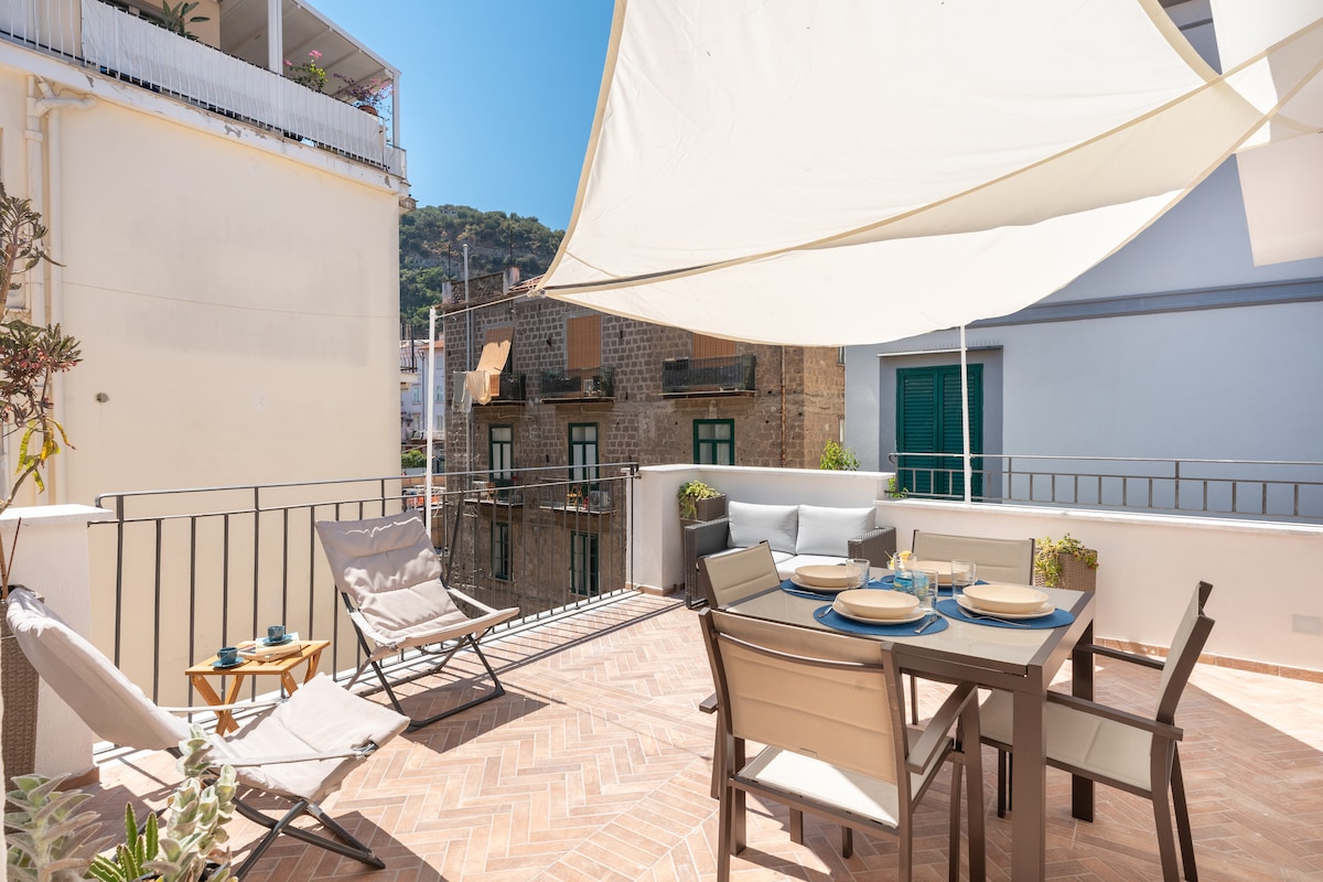An outdoor terrace offers a dining table set for four, surrounded by comfortable chairs and loungers. A large umbrella provides shade, and potted plants add greenery. The scenic backdrop includes nearby buildings and a hillside, enhancing the inviting space for alfresco dining and relaxation.