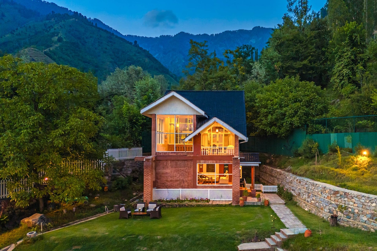 A charming homestay is nestled among lush greenery, illuminated by warm indoor lighting. The two-story structure features expansive windows showcasing the inviting interior. A spacious lawn, bordered by stone pathways and adorned with seating, complements the scenic mountain backdrop.