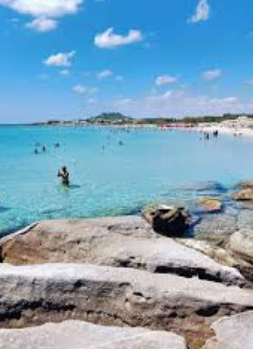 A tranquil beach scene is depicted with clear turquoise waters lapping at smooth, sandy shores. Sunbathers and swimmers enjoy the warm weather, while rocky formations frame the water's edge. Gentle waves create a serene atmosphere under a bright blue sky dotted with fluffy white clouds.