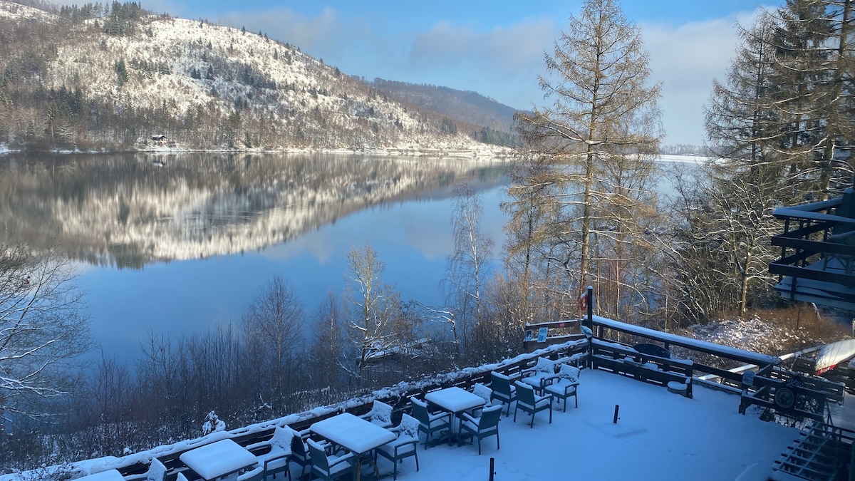 A serene landscape is depicted, showcasing a tranquil lake bordered by snow-covered hills. The clear water reflects the surrounding trees and sky, creating a harmonious natural scene. A deck with several tables is visible in the foreground, offering picturesque views of the winter scenery.
