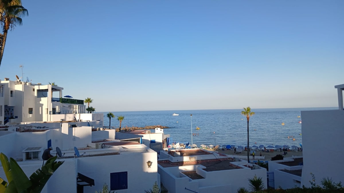 A serene beachfront view captures the tranquil sea and sandy shore. White buildings with blue accents frame the scene, while palm trees add a touch of greenery. The horizon blends softly with the sky, suggesting a calm and inviting atmosphere.