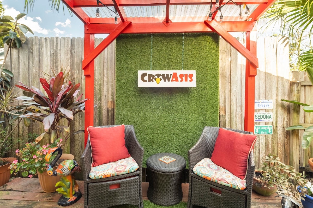 A colorful outdoor seating area features two wicker chairs with patterned cushions, positioned on a wooden deck. A round table sits between them, and vibrant green plants surround the space. The CrowAsis sign is mounted on a textured green wall, enhancing the charming ambiance.
