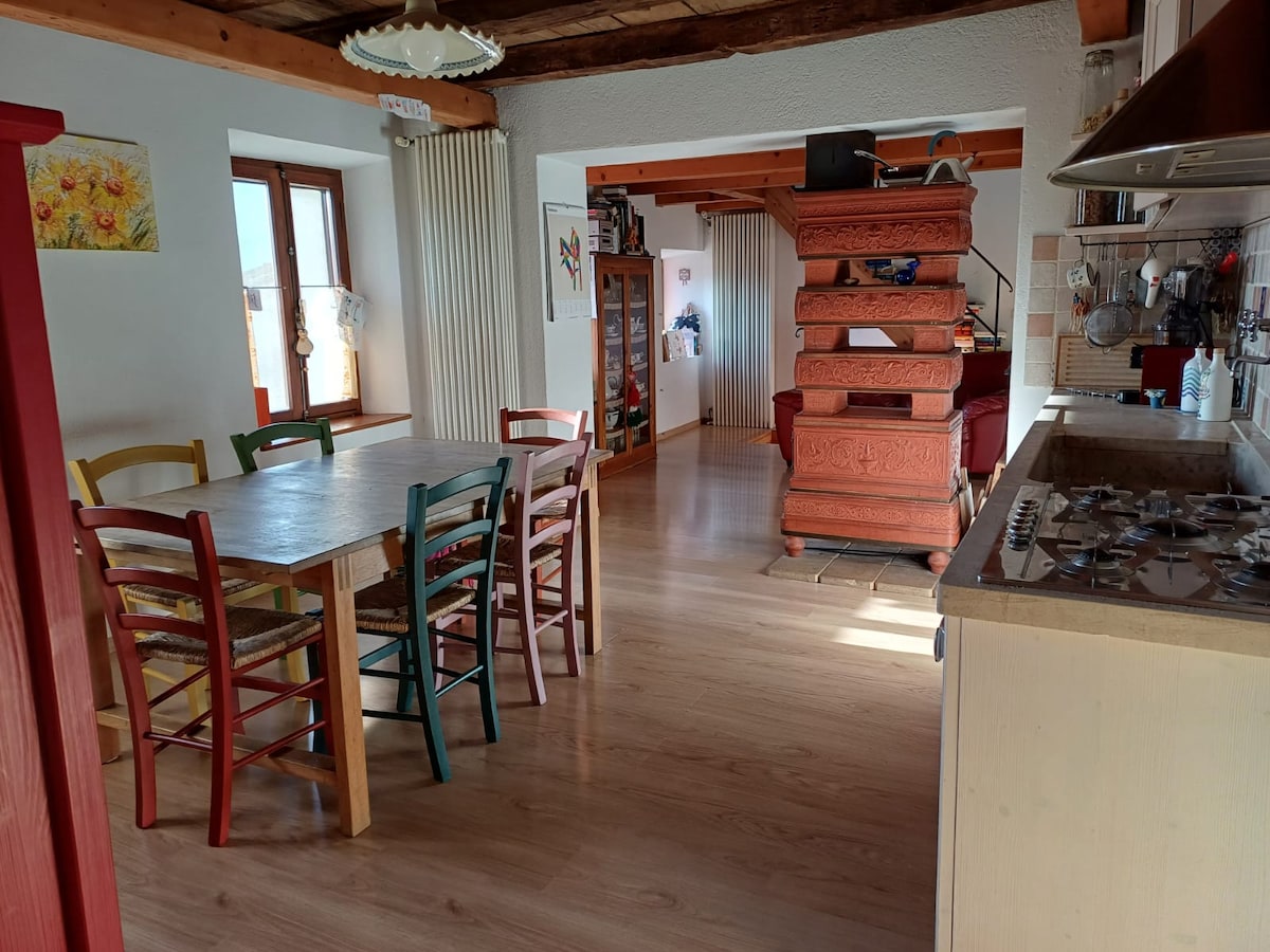 A spacious dining area features a wooden table surrounded by six colorful chairs. Natural light enters through two windows, illuminating the wooden beams of the ceiling. In the background, a staircase leads to the upper level, and a kitchen area with a stove is visible.