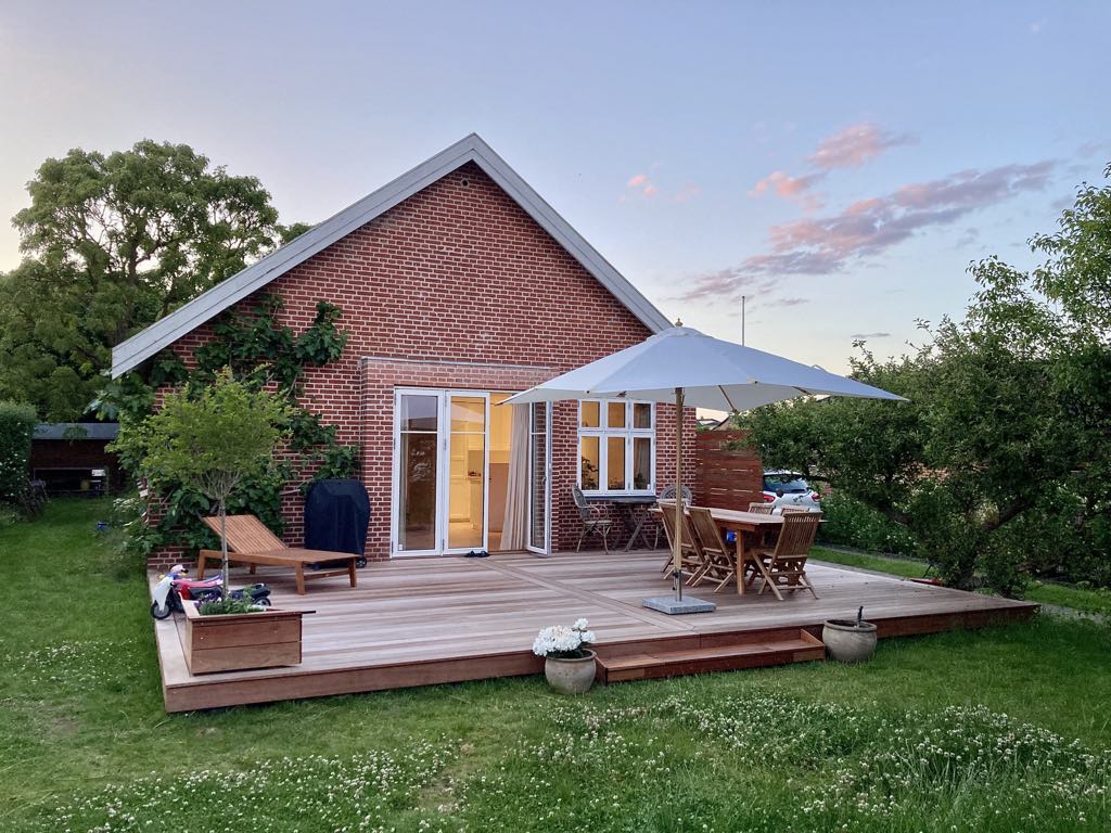 A charming wooden terrace features a large umbrella shading a dining table and lounge chairs. The brick house, surrounded by greenery, showcases large glass doors that connect the interior to the outdoor space, creating a welcoming atmosphere for al fresco dining.