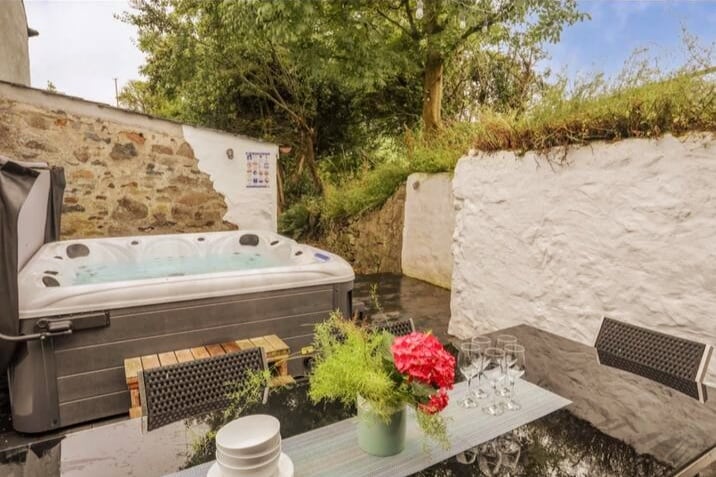 An outdoor area features a hot tub positioned on a wooden deck, surrounded by rustic walls. A glass-top table is set nearby, adorned with a vibrant floral centerpiece, and glassware is neatly arranged, set against the backdrop of lush greenery.