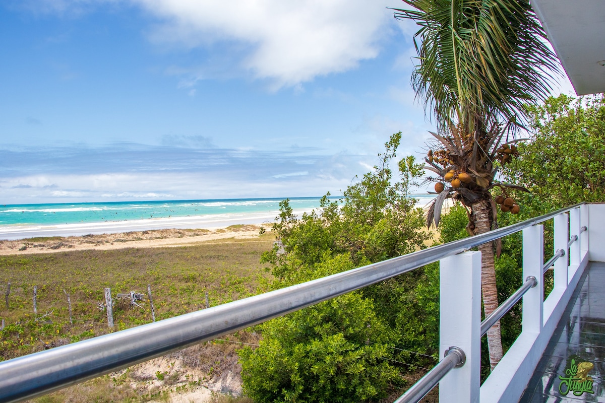 A balcony view showcases a serene beach bordered by vibrant greenery and palm trees. The landscape features gentle waves lapping against the shore, with a clear blue sky above, creating a tranquil atmosphere ideal for relaxation.