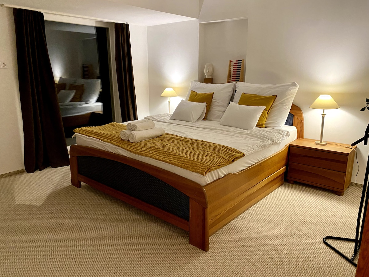 A well-appointed bedroom features a spacious bed with plush white linens and decorative yellow pillows. Bedside tables with lamps provide soft lighting, complementing the neutral color scheme. A large window is framed by dark curtains, allowing for a serene and calming atmosphere.