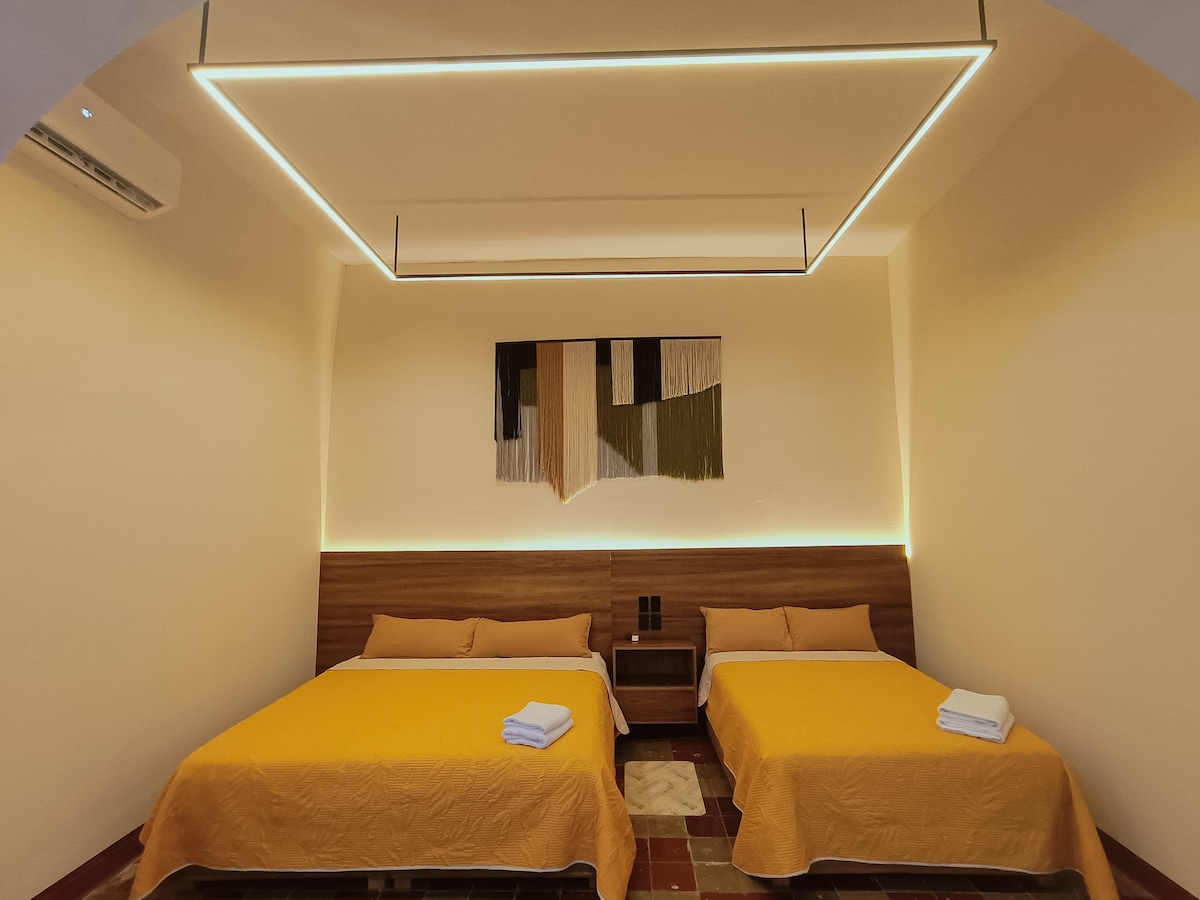 A spacious sleeping area is equipped with two beds covered by golden-yellow bedspreads. White towels are neatly placed at the foot of each bed. The room features soft lighting and a tapestry artwork is displayed above the headboard, enhancing the contemporary design.