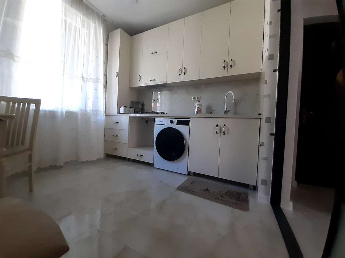 The kitchen area is equipped with light-colored cabinets and a washing machine. A small table and chair are positioned near the window, allowing natural light to fill the space. A subtle area rug is located in front of the washing machine.